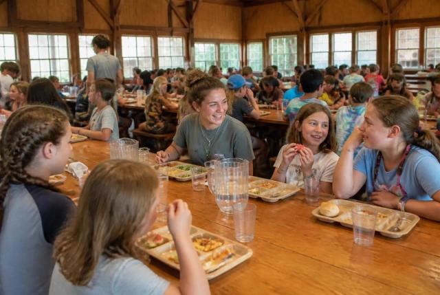 Campers and counselors eating together at long wooden tables in a camp dining hall